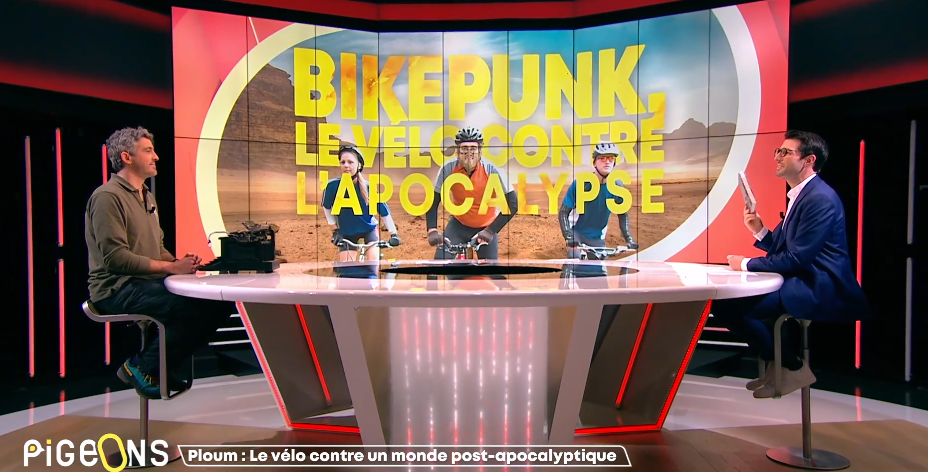 Ploum sur le plateau de l’émission télé On n’est pas des pigeons avec le présentateur Simon François. En arrière plan, un écran annonce Bikepunk, le vélo contre l’apocalypse